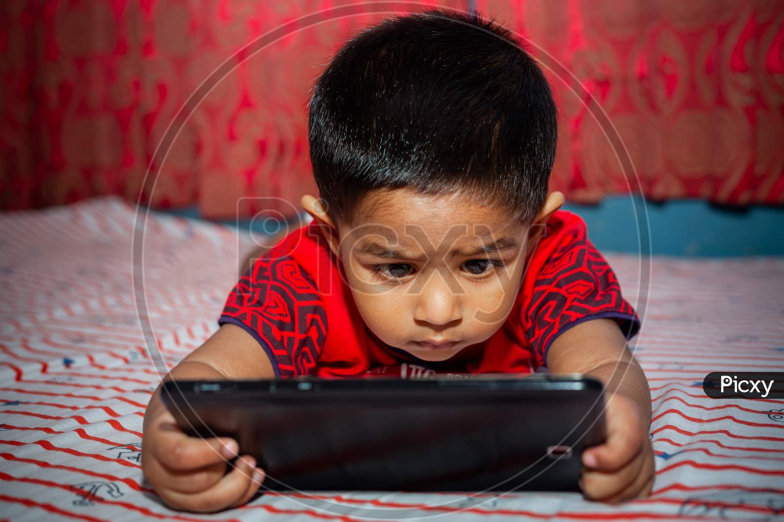 A Child With Full Attention On His Bed Watching Cartoons Using The Tab Smartphone. Kids Playing With Smartphone. Mobile Phone And Internet Addiction Concept.