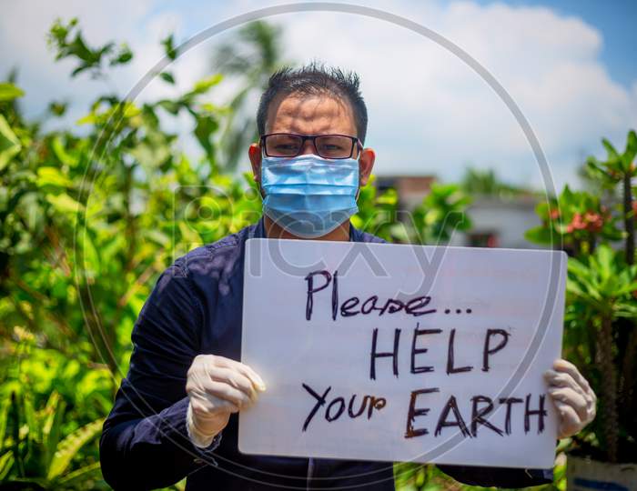 A Young Man Wearing A Medical Mask And Safety Gloves Sitting With A Placard Message To "Please Help Your Earth". A Man Holding A Placard Message In Outdoor Natural Light.