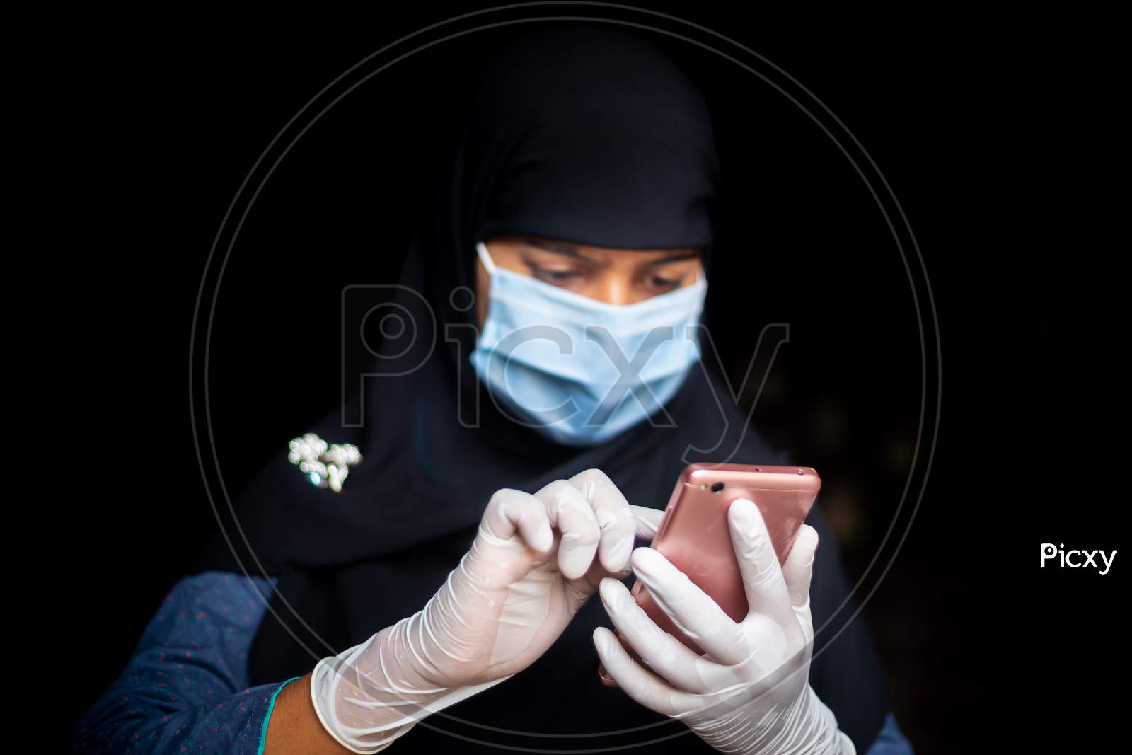 A Safety Mask And Gloves-Wearing A Muslim Girl Is Browsing His Smartphone For Coronavirus News. Black Hijab Woman Wearing A Blue Mask For Coronavirus Safety.