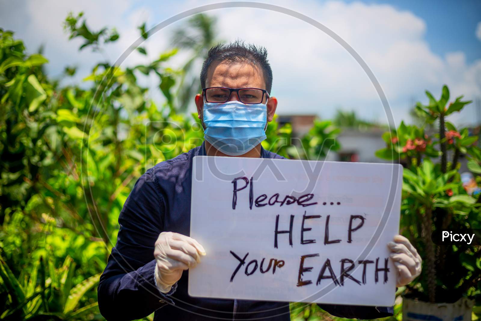 A Young Man Wearing A Medical Mask And Safety Gloves Sitting With A Placard Message To "Please Help Your Earth". A Man Holding A Placard Message In Outdoor Natural Light.