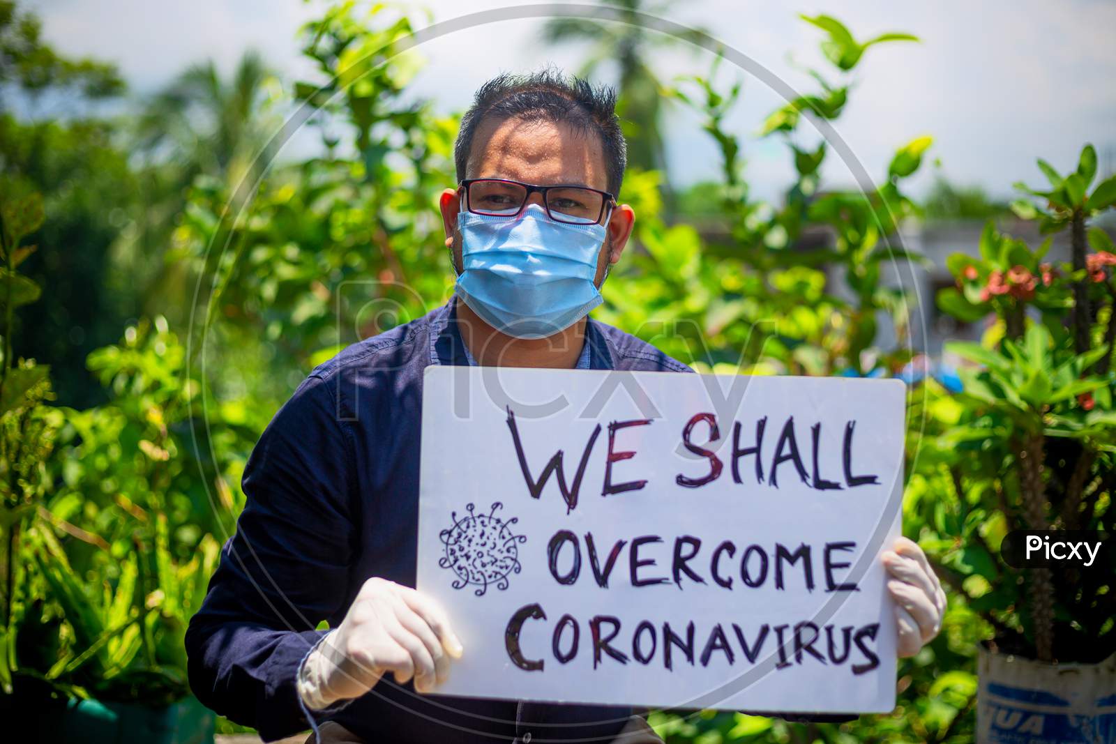 A Young Man Wearing A Medical Mask And Safety Gloves Stands With A Placard Message To "We Shall Overcome Coronavirus".A Man Holding A Placard Message In Outdoor Natural Light.