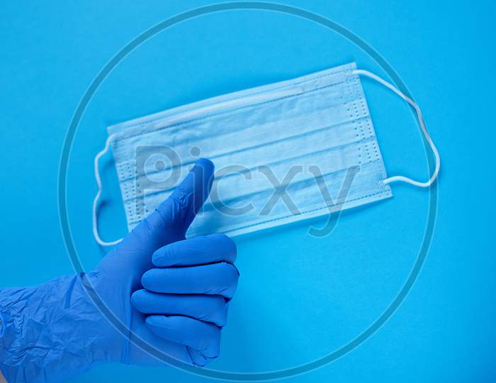 A Hand In Medical Glove Thumb Up Over Protective Mask. Typical Surgical Mask For Covering The Mouth And Nose. Protect From Coronavirus And Bacteria Concept On Blue Background