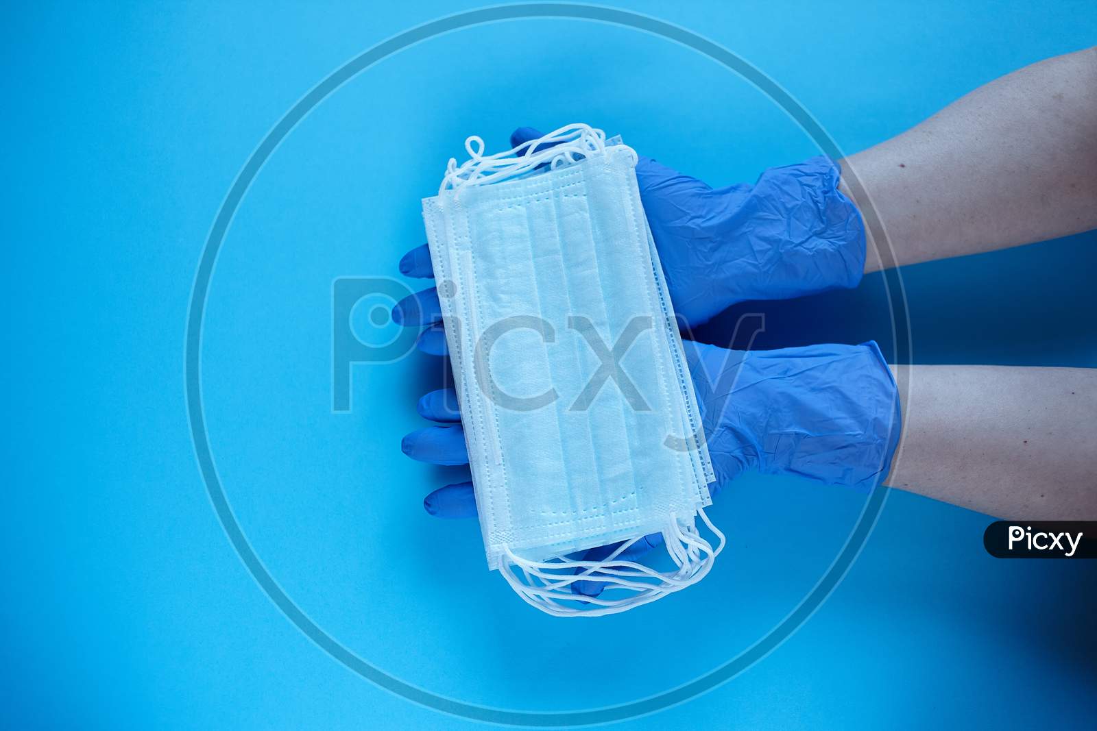 Two Hands Of A Doctor In Medical Gloves Holds, Takes, Gives Medical Surgical Masks For Covering The Mouth And Nose. Top View, Close Up, On Blue Background