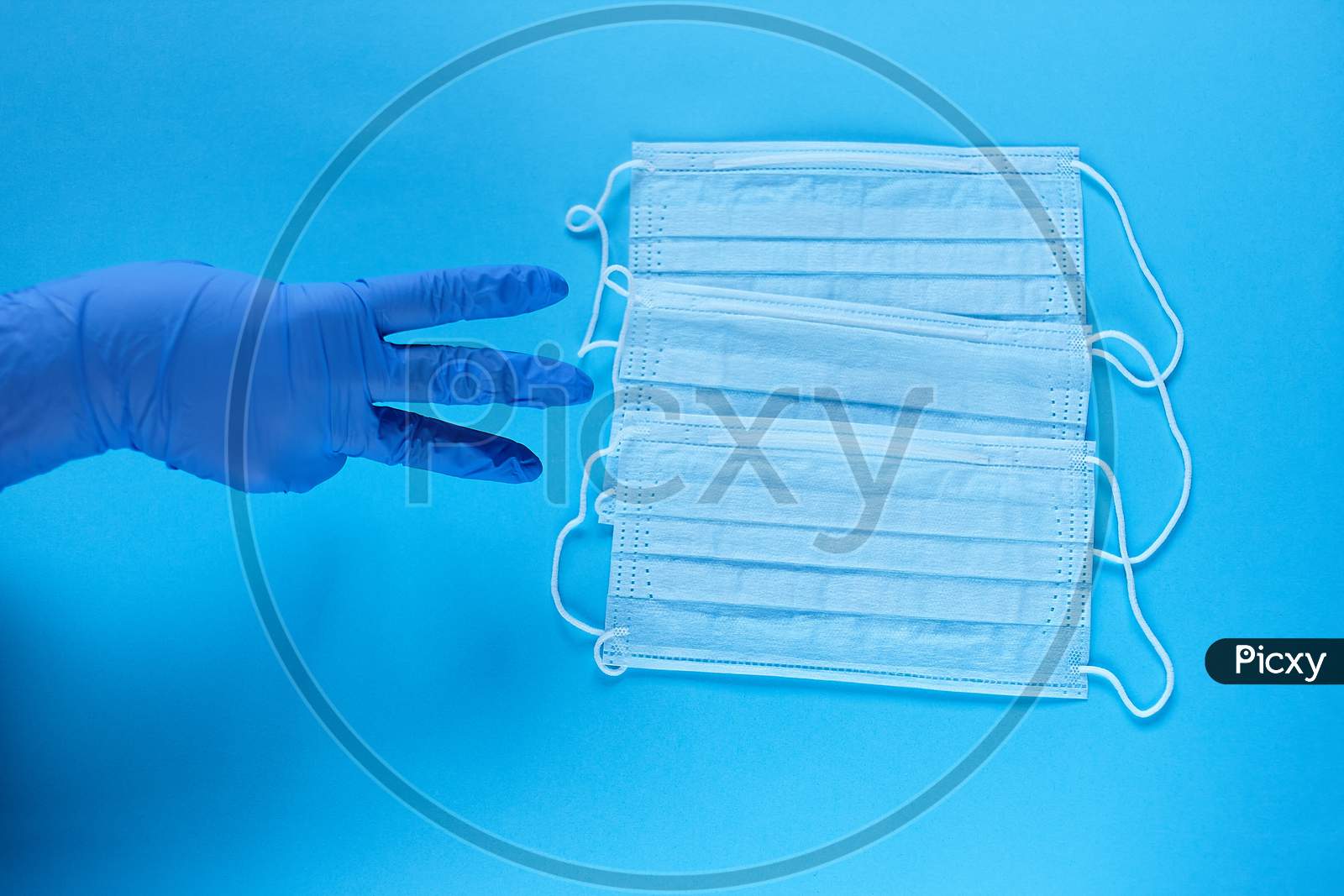 A Hand In Protective Medical Glove Points A Fingers At Three Protective Medical Masks. Protect From Coronavirus For Three People Concept On Blue Background