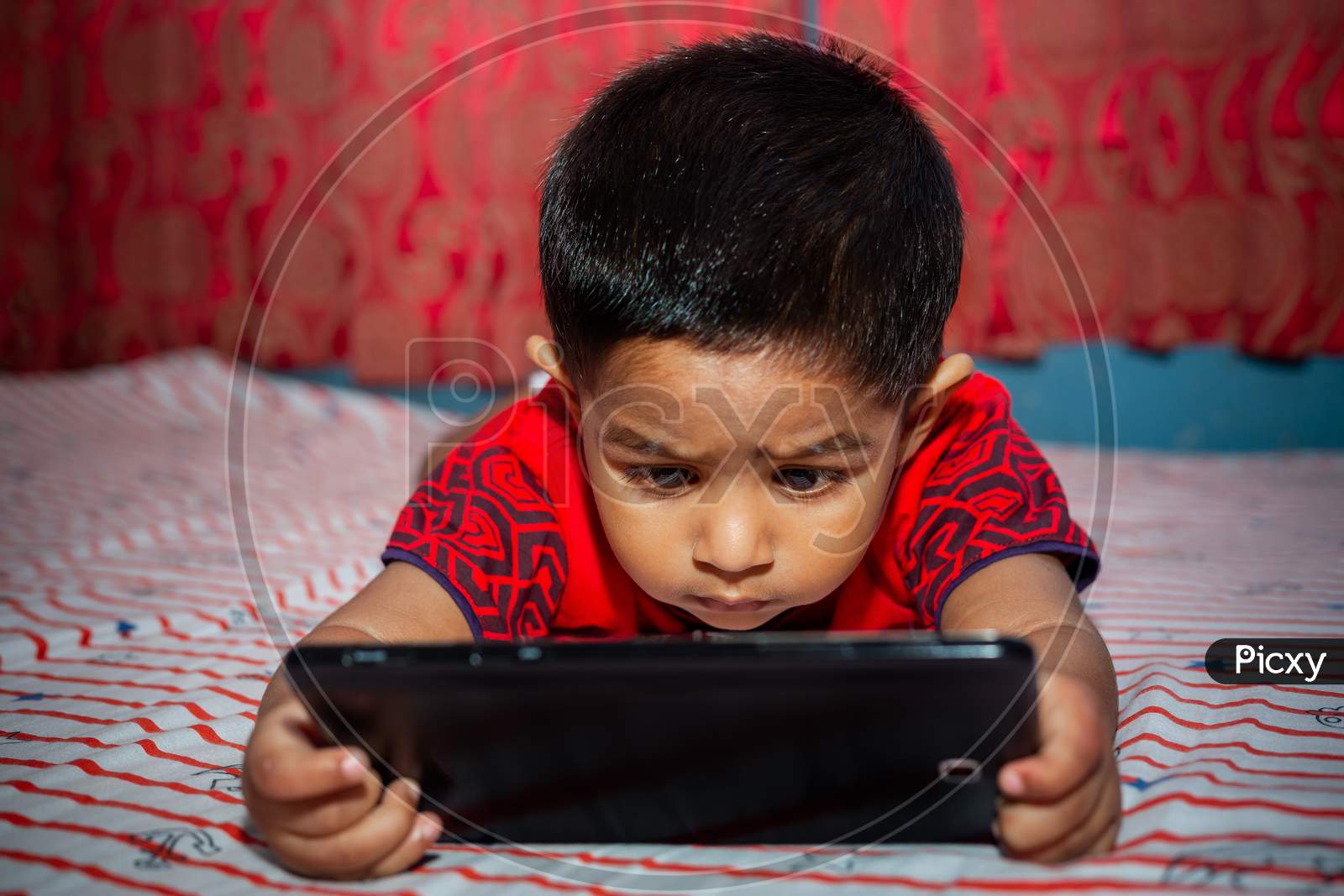 A Child With Full Attention On His Bed Watching Cartoons Using The Tab Smartphone. Kids Playing With Smartphone. Mobile Phone And Internet Addiction Concept.