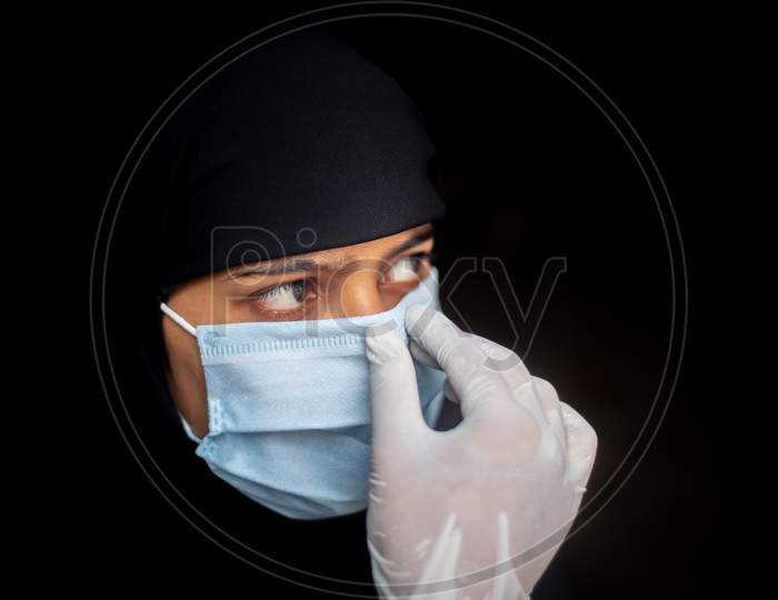 Muslim Girl Adjusting A Surgical Mask For Coronavirus Protection. Black Hijab Woman Wearing A Blue Mask For Safety. Dark Background Views.