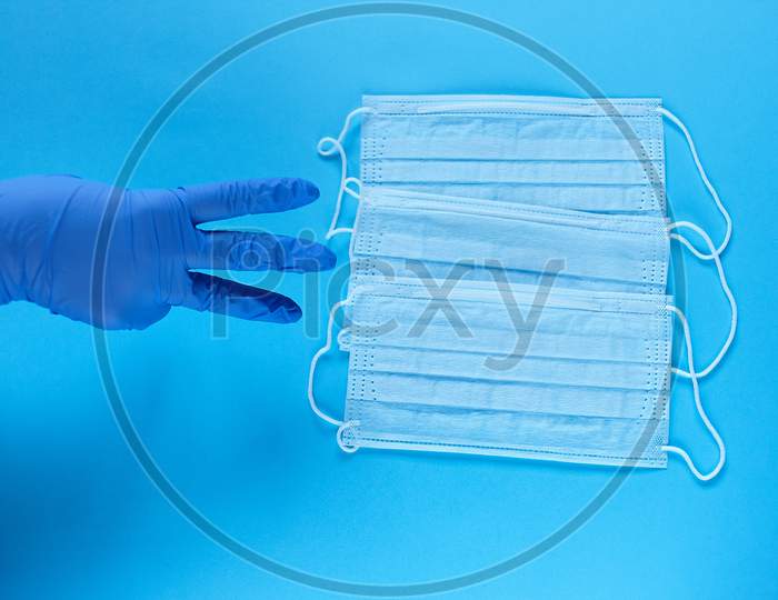 A Hand In Protective Medical Glove Points A Fingers At Three Protective Medical Masks. Protect From Coronavirus For Three People Concept On Blue Background