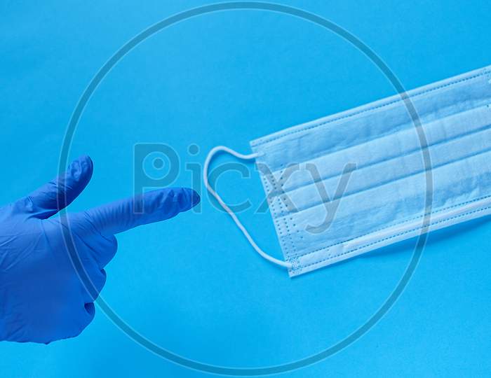 A Hand In A Protective Medical Glove Points A Finger At A Protective Medical Mask. Protect From Coronavirus Concept On Blue Background