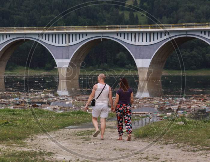 Couple Walking Next To Polluted Lake Near Bridge
