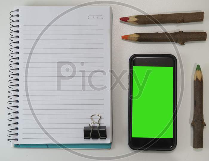 Top View Of Office Desk With Blank Note Book, Rustic Colored Pencils And Smartphone.