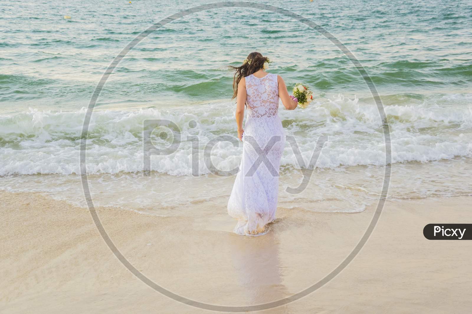 Back View Of Bride Entering The Sea With Wedding Dress And Bouquet In Hand.