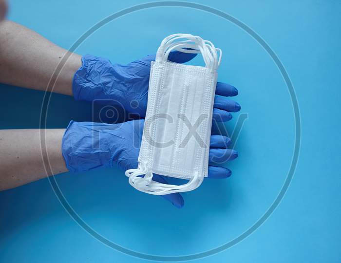 Two Hands Of A Doctor In Medical Gloves Holds, Takes, Gives Medical Surgical Masks For Covering The Mouth And Nose. Top View, Close Up, On Blue Background
