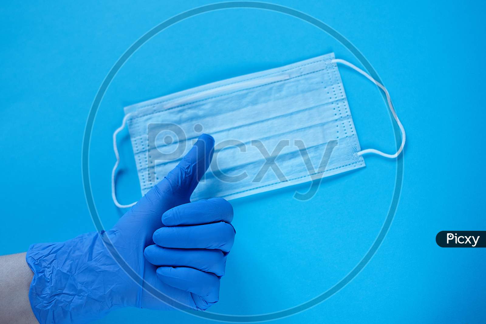 A Hand In Medical Glove Thumb Up Over Protective Mask. Typical Surgical Mask For Covering The Mouth And Nose. Protect From Coronavirus And Bacteria Concept On Blue Background
