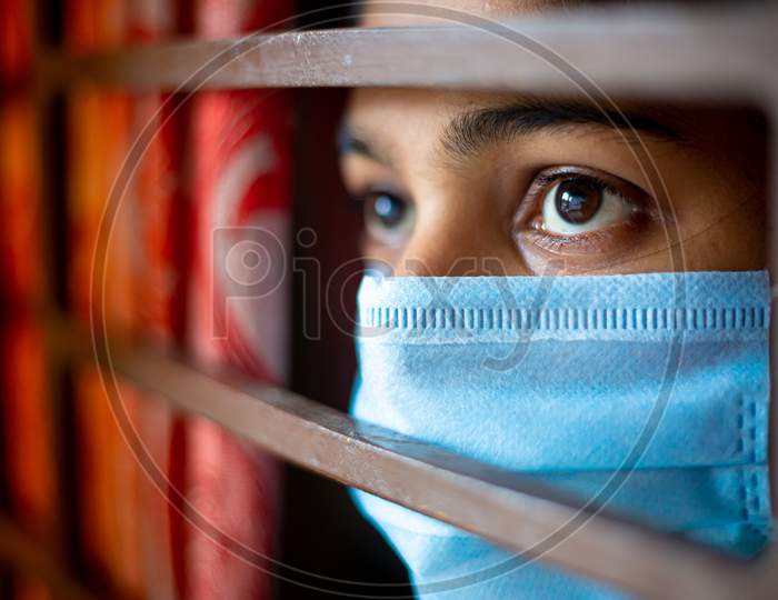 A Bored Asian Young Girl Wearing A Protection Surgical Face Mask At Looking Through Window Being In-Home Quarantine During Coronavirus Outbreak. Close-Up Views.