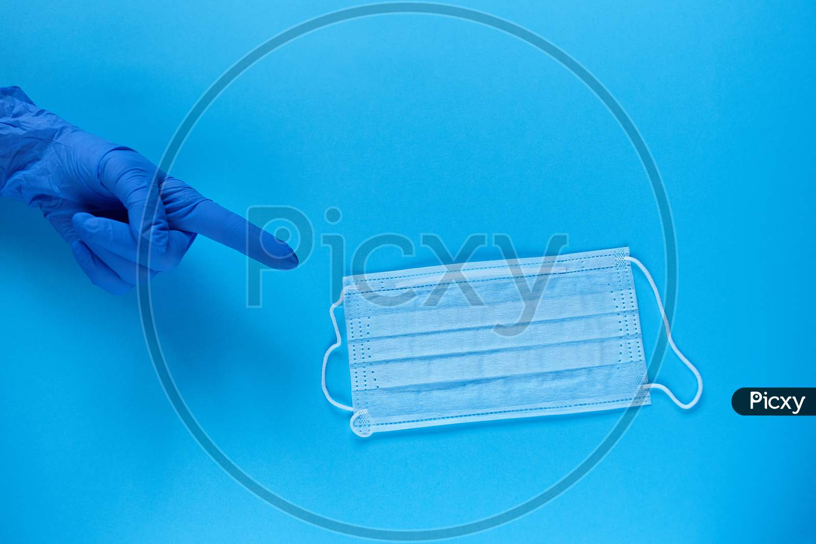 A Hand In A Protective Medical Glove Points A Finger At A Protective Medical Mask. Typical Surgical Mask For Covering The Mouth And Nose. Protect From Coronavirus And Bacteria Concept On Blue Background