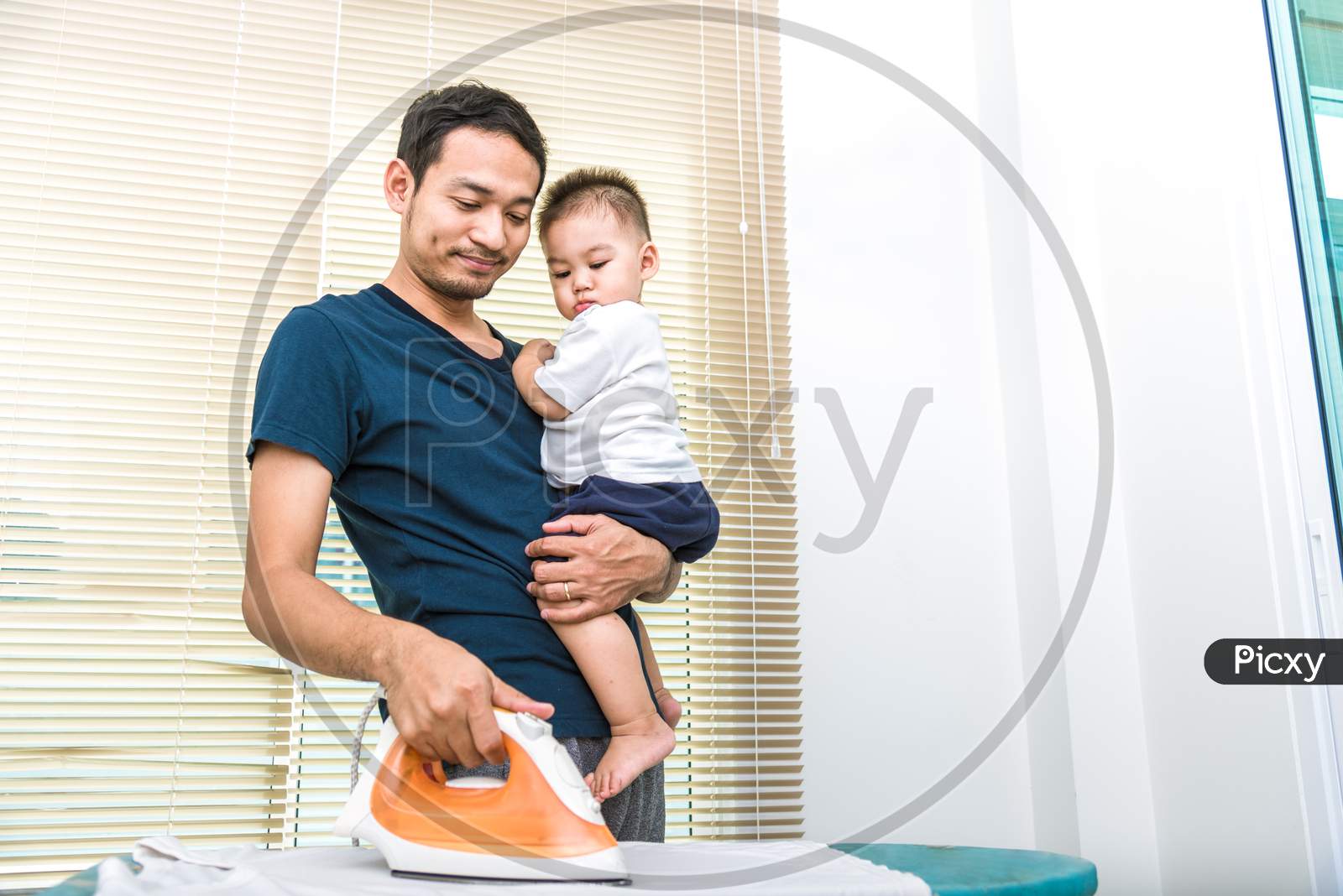 Single Dad Is Ironing While Carrying His Son. People And Lifestyles Concept.