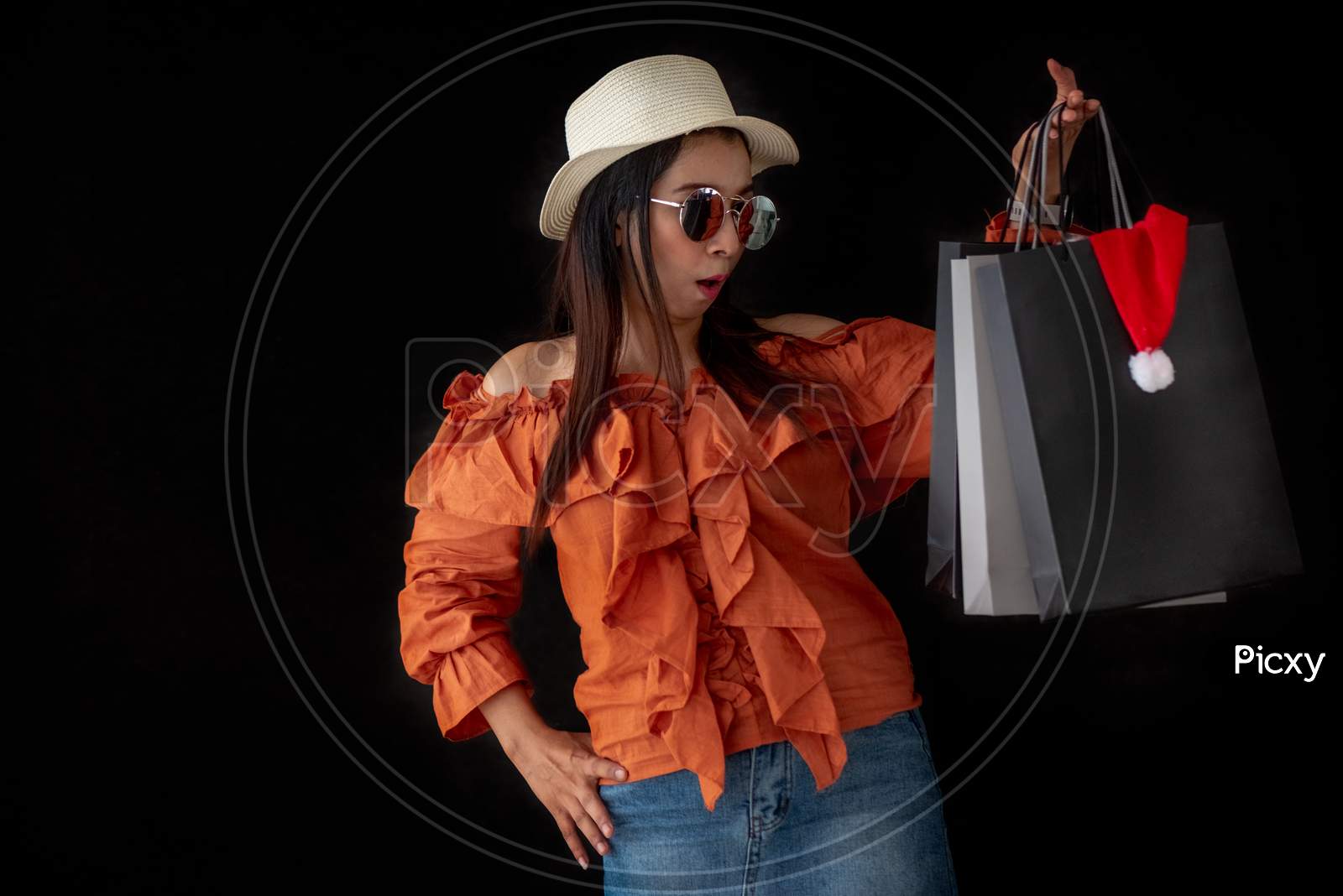 Asian Shopping Woman Surprised With Black Friday Shopping Bag And Santa Claus Hat Inside On Black Background. Shopaholics And Beauty Fashion Theme.