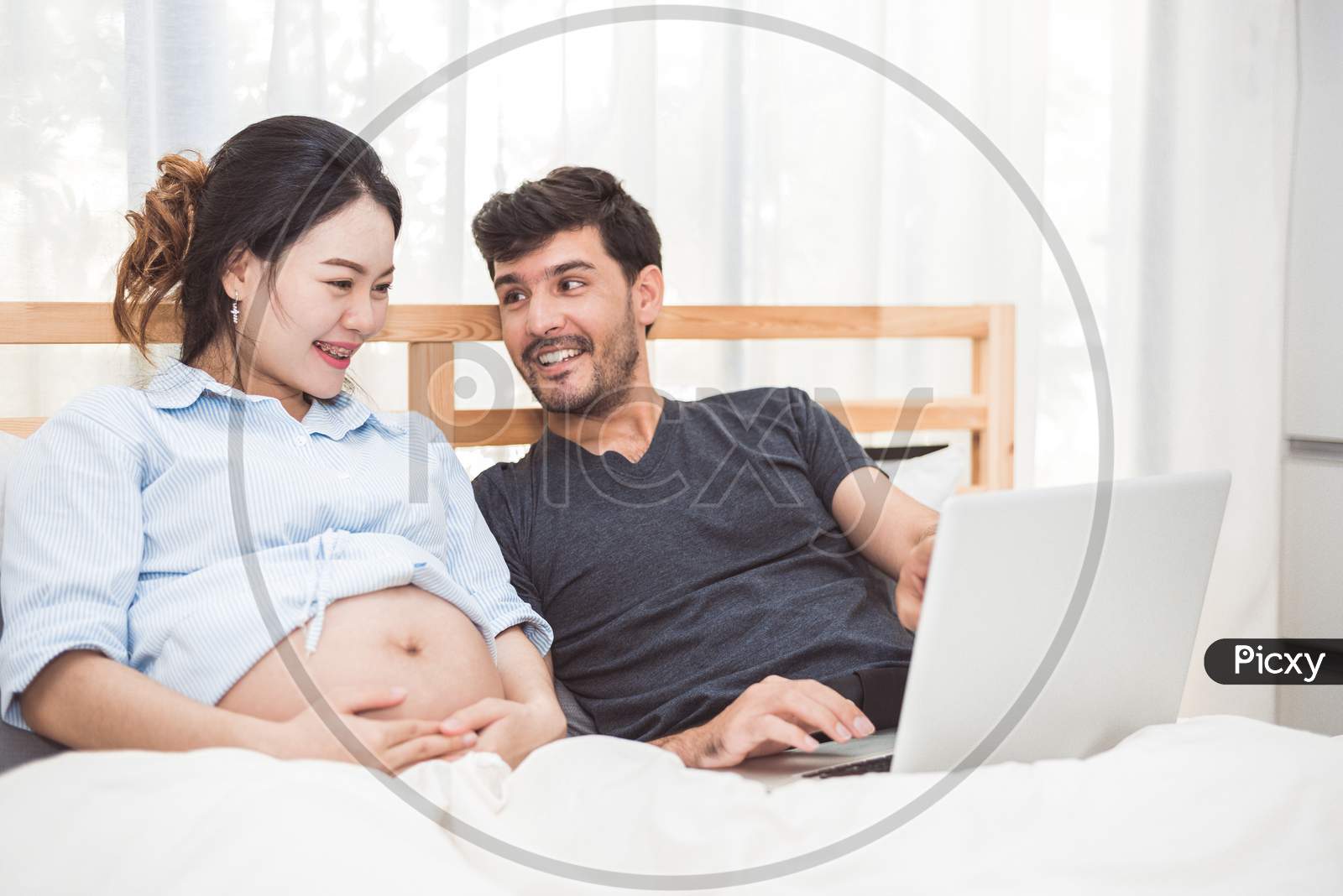 Happy Man And His Pregnant Wife Using Laptop To Searching Newborn Baby Items For Preparing Parenthood. Couple Lifestyle Family And Technology Concept. Women Health And Medical Theme.