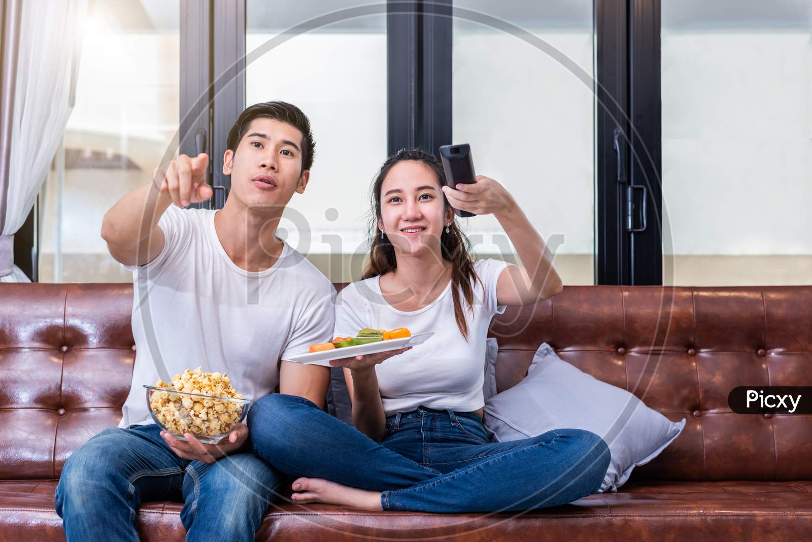 Asian Couples Watching Television Together On Sofa In Their Home. People And Lifestyles Concept. Vacation And Holiday Concept. Honeymoon And Pre Wedding Theme. Happy Family Activity Theme