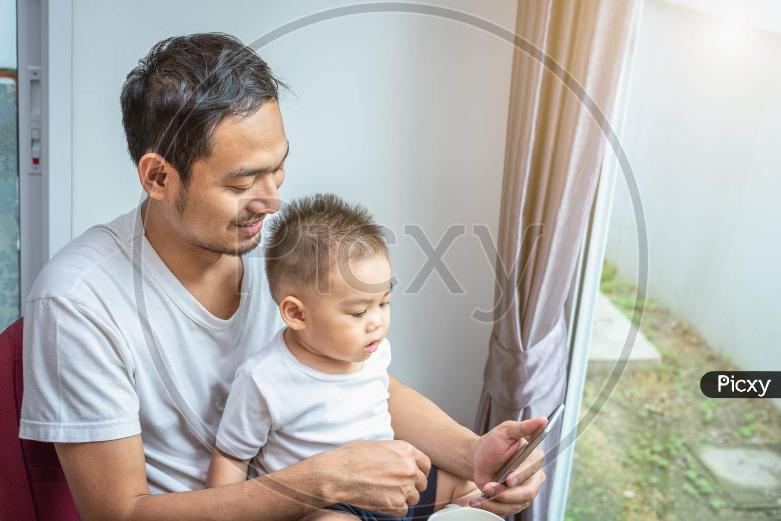 Asian Father And Son Using Smart Phone Together In Home Background. Technology And People Concept. Lifestyles And Happy Family Theme. Internet And Communication Theme
