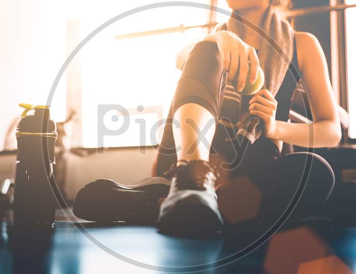 Sport Woman Sitting And Resting After Workout Or Exercise In Fitness Gym With Protein Shake Or Drinking Water On Floor. Relax Concept. Strength Training And Body Build Up Theme. Warm And Cool Tone