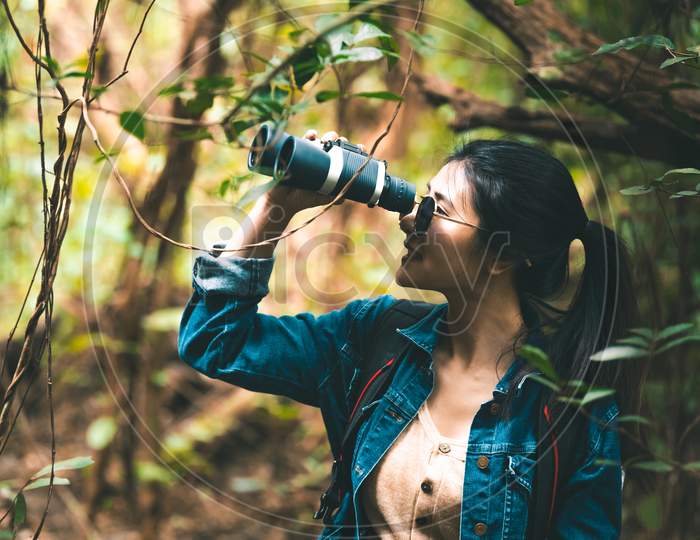 Beautiful Asian Woman With Binoculars Telescope In Forest Looking Destination. People Lifestyles And Leisure Activity. Nature And Backpacker Traveling Jungle Background. Bird Watching And Living