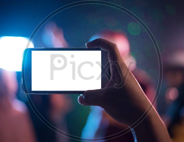 Hand Holding Smart Phone With Blank White Screen. Photo Shot Blurry Party And Concert Of Event Background And Copy Space. Smart Phone White Screen. Technology And Lifestyle