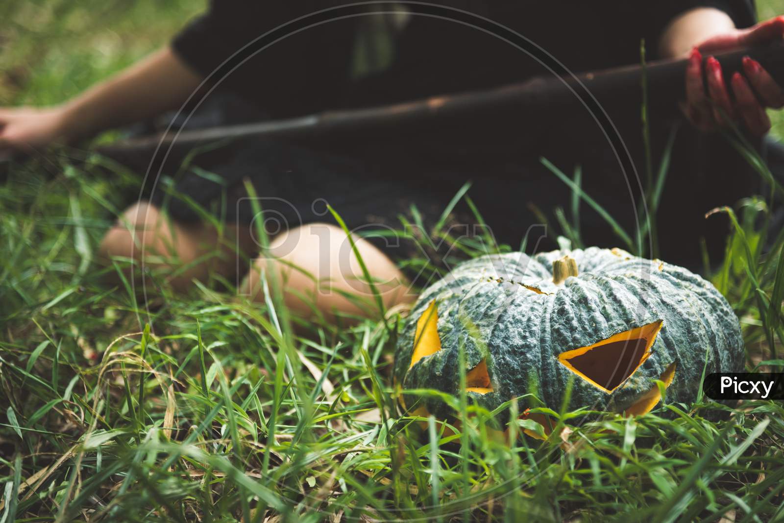Close Up Of Green Pumpkin Lantern On Grass With Female Wizard Holding Magic Wand Background. Halloween Day And Horror Concept. Cosplay And Playful Theme. Nature And Decoration Theme.