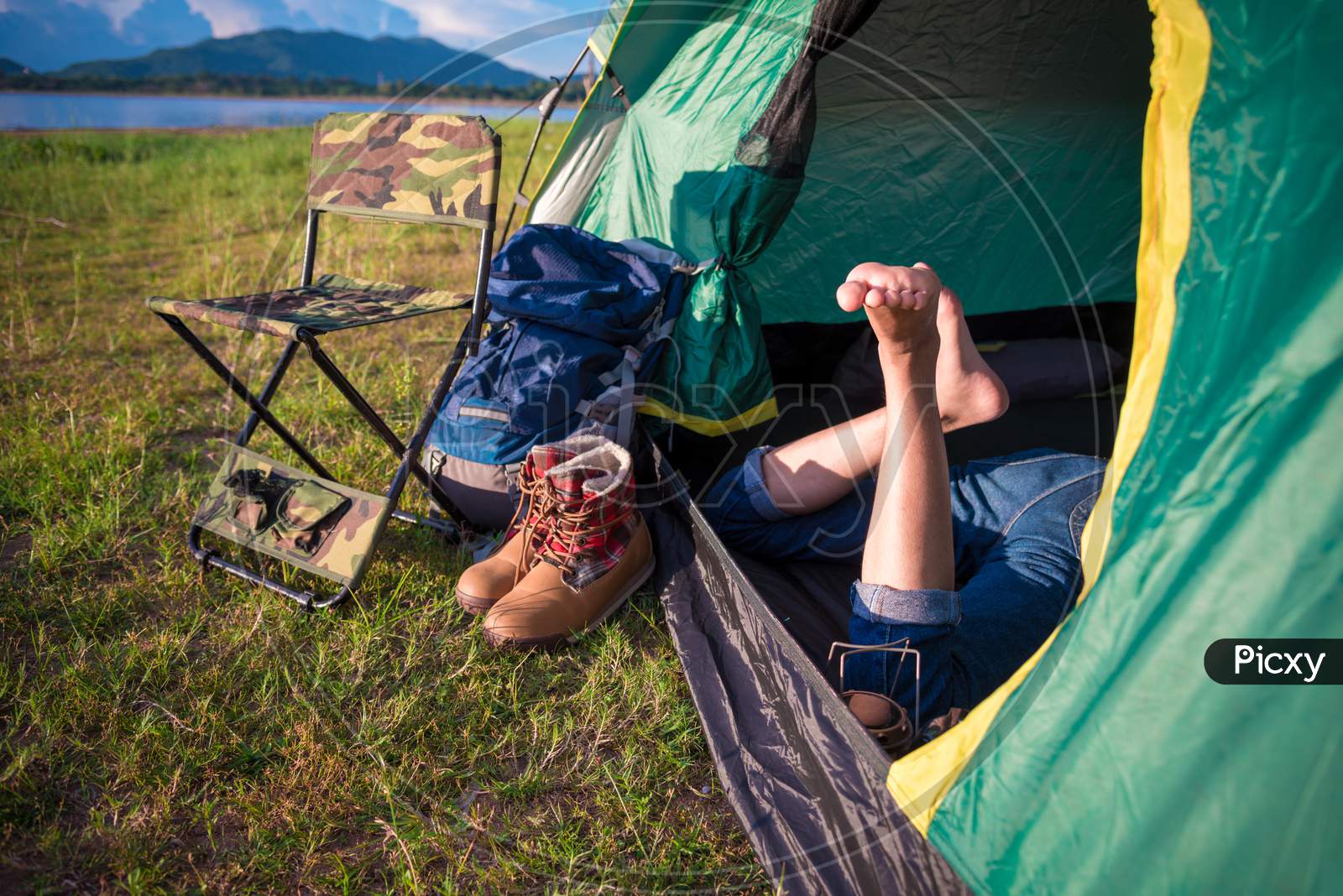 Close Up Of Woman Legs Relaxing In Camping Tent With Mountain Lake And Meadow And Grass Field Background. Lifestyles And People Concept. Camping And Picnec Theme. Green Natural And Summer Travel Theme