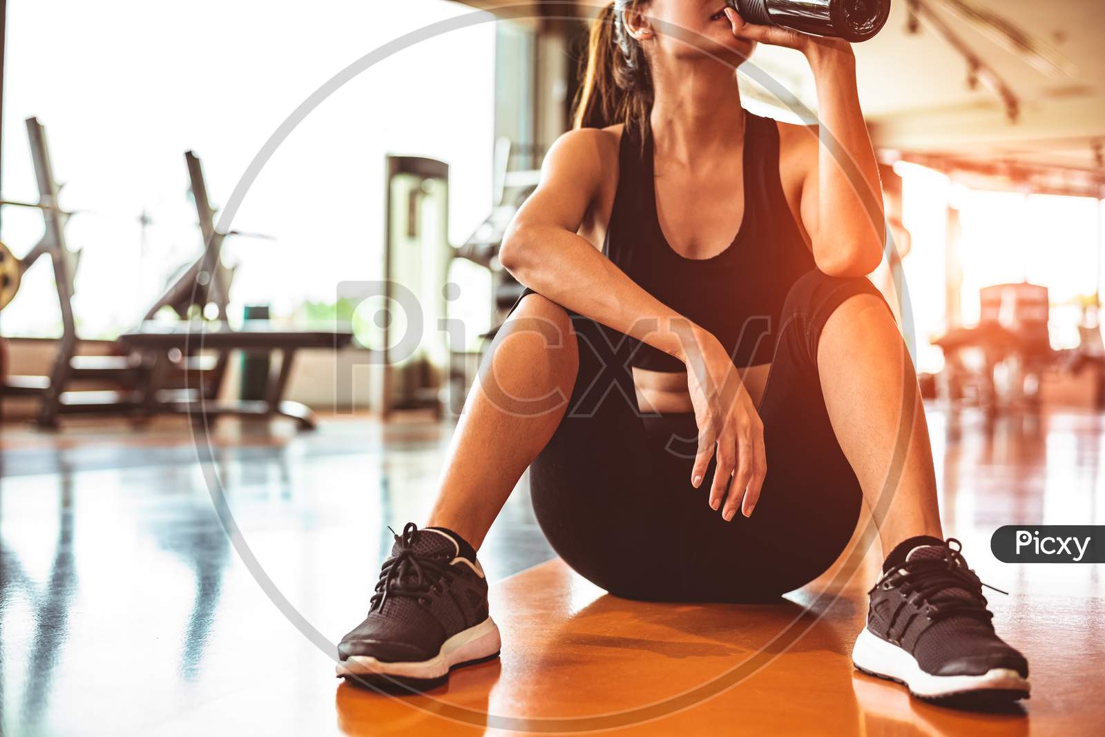 Sport Woman Relax Resting After Workout Or Exercise In Fitness Gym. Sitting And Drinking Protein Shake Or Drinking Water On Floor. Strength Training And Bodybuilder Muscle Theme. Warm And Cool Tone