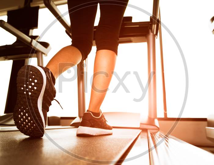 Back View Of Lower Body Legs Of Fitness Girl Running On Machine Or Treadmill In Fitness Gym With Sun Ray. Warm Tone. Healthy And Exercise Activity Concept. Workout And  Strength Training Theme.