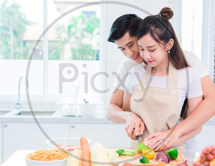 Asian Lovers Or Couple Cooking And Slicing Vegetable In Kitchen Room. Man And Woman Looking Each Other In Home. Holiday And Honeymoon Concept. Valentine Day And Wedding Theme