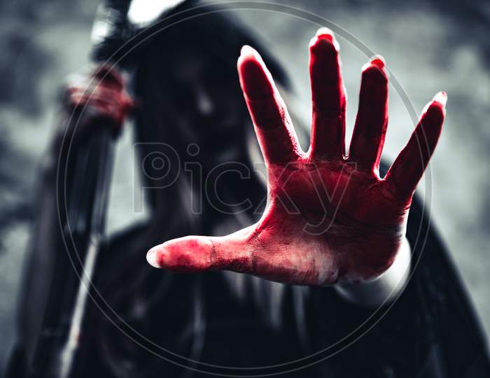 Witch Showing Bloody Hand With Reaper. Female Demon Angel In Black Clothes And Hood On Grunge Wall Background. Halloween Day And Mystery Concept. Fantasy Of Magic Theme. Afterlife And Death Concept.