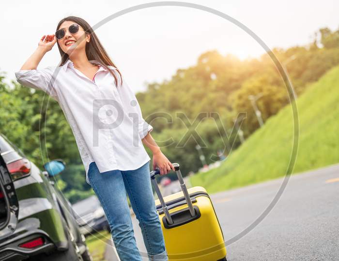 Beauty Asian Woman With Sunglasses Dragging Yellow Suitcase Baggage Alone On Road Trip. People Lifestyle And Vacation Concept. Nature And Summer Background. Girl Having Car Adventure Transportation