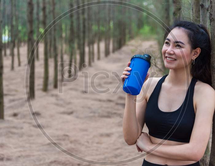 Beauty Asian Sport Woman Resting And Holding Drinking Water Bottle And Relaxing In Middle Of Forest After Tired From Jogging. Girl Looking Attraction View. Workout Concept. Lifestyle Theme