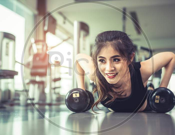 Asian Young Woman Doing Push Ups With Dumbbell On Floor In Fitness Gym And Equipment Background. Workout And Sport Exercise Concept. Healthy And Happiness Concept. Beauty And Body Build Up Theme.
