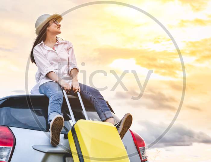 Happy Asian Woman On Top Of Car With Luggage Bag. Girl Sitting On Roof And Looking Sunset Before Night In Evening. People Lifestyle In Long Vacation Trip Concept. Nature And Transportation Vehicle.