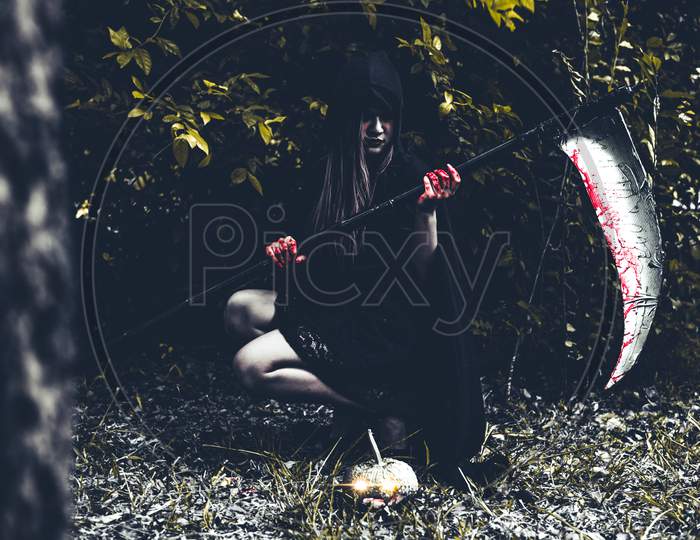 Demon Witch Sitting And Holding Reaper In Black Forest. Halloween Day Concept.