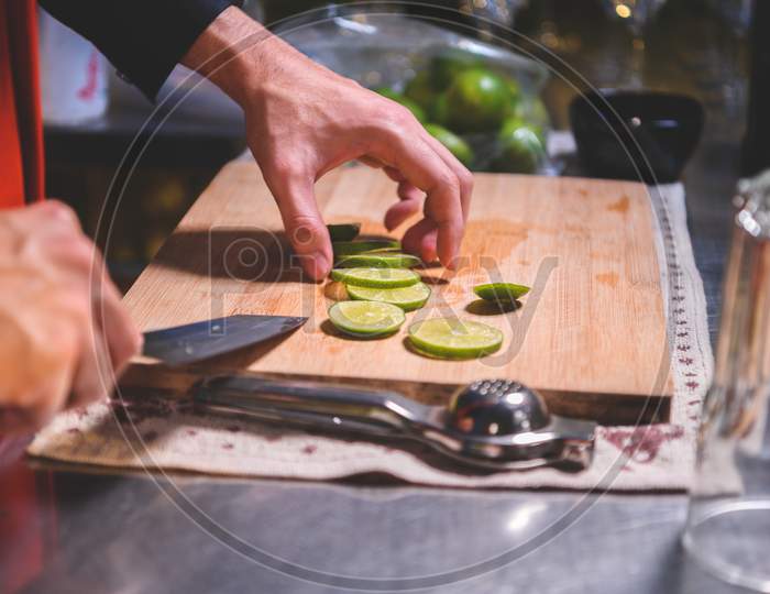Closeup Of Professional Bartender Hand Slicing Lemon For Making Lemonade Juice By Knife In Night Club. Chef Making Drinks For Guest In Pub Restaurant. Food And Beverage Concept.