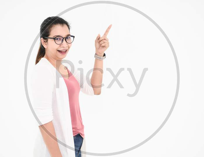 Asian Woman Posing And Pointing Finger At Blank Space With Casual Outfit And Eyeglasses In Happy Mood On White Isolated Background. People Beauty And Portrait Concept. Teacher Or Professor Acting.