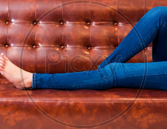 Woman Legs With Wearing Jeans While Relaxing At Movie Theater Or Home On Brown Leather Sofa, Relax And Holiday Concept, Bed Time Sleeping Concept. People Lifestyle And Holiday. Closeup Girl Foot