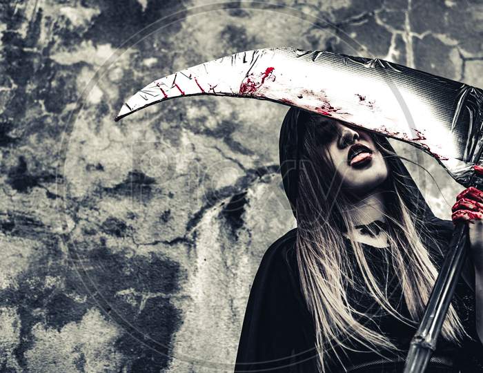 Demon Witch Licking Blood On Reaper. Female Demon Angel In Black Clothes And Hood On Grunge Wall Background. Halloween Day And Mystery Concept. Fantasy Of Magic Theme. Afterlife And Death Concept.