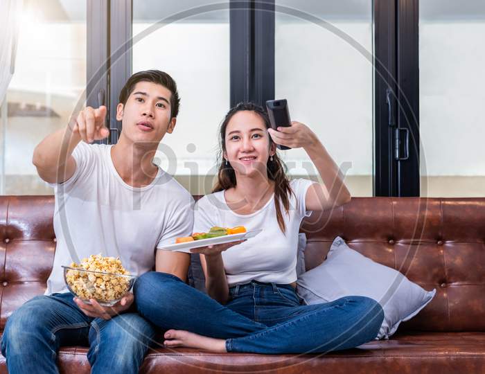 Asian Couples Watching Television Together On Sofa In Their Home. People And Lifestyles Concept. Vacation And Holiday Concept. Honeymoon And Pre Wedding Theme. Happy Family Activity Theme