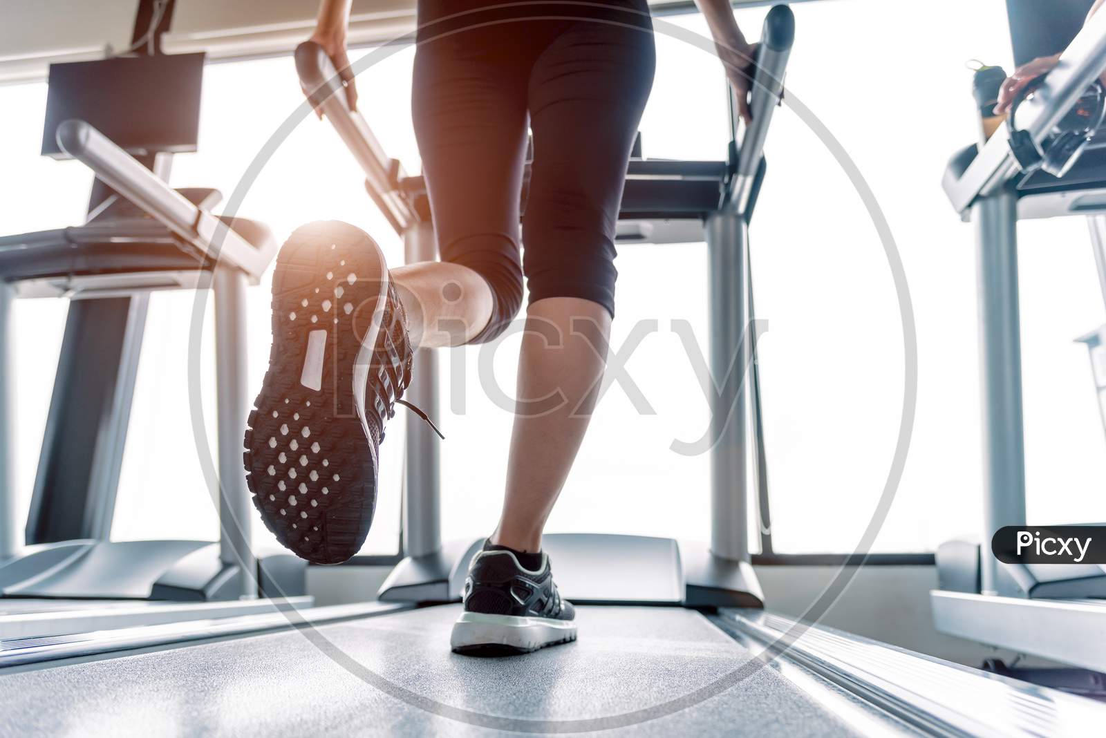 Lower Body At Legs Part Of Fitness Girl Running On Running Machine Or Treadmill In Fitness Gym With Sun Ray. Warm Tone. Healthy And Exercise Activity Concept. Workout And  Strength Training Theme.