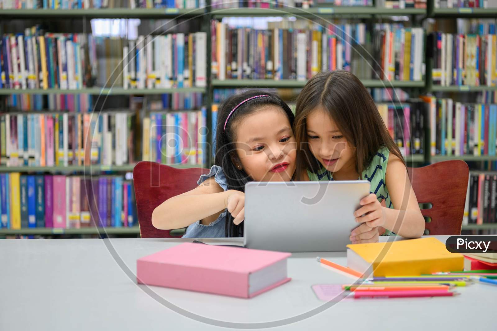 Two Little Happy Cute Girls Playing On A Tablet Pc Computing Device In Library At School. Education And Self Learning Wireless Technology Concept. People Lifestyles And Friendship. Preschool Children