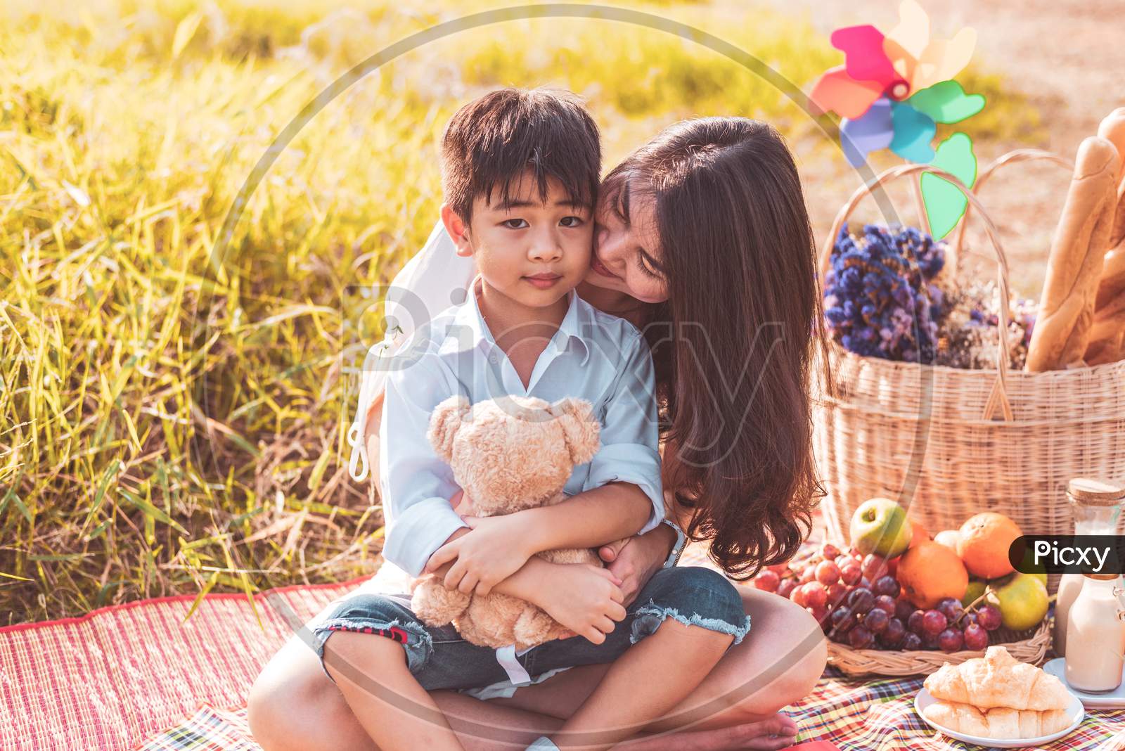 Little Asian Boy Kissed At Cheek By His Mom In Meadow When Doing Picnic. Mother And Son Playing Together. Celebrating In Mother Day And Appreciating Concept. Summer People And Lifestyle Theme.