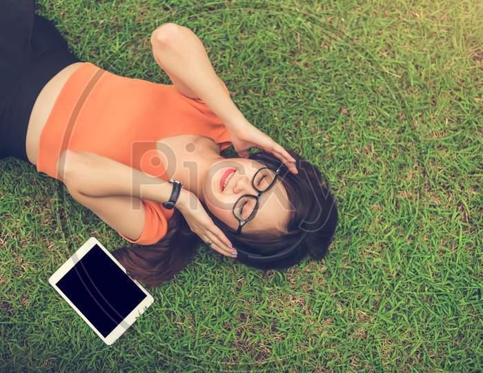 Asian Woman Listening Music With Blank Tablet Black Isolated Screen On Grass In Park. People Lifestyle And Entertainment Concept.