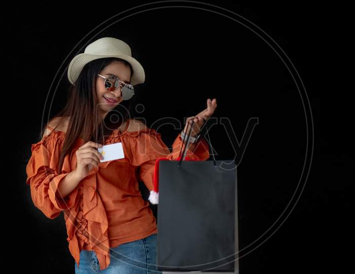Asian Shopping Woman Holding Credit Card With Black Friday Shopping Bag And Santa Claus Hat Inside On Black Background. Shopaholics And Beauty Fashion Theme.