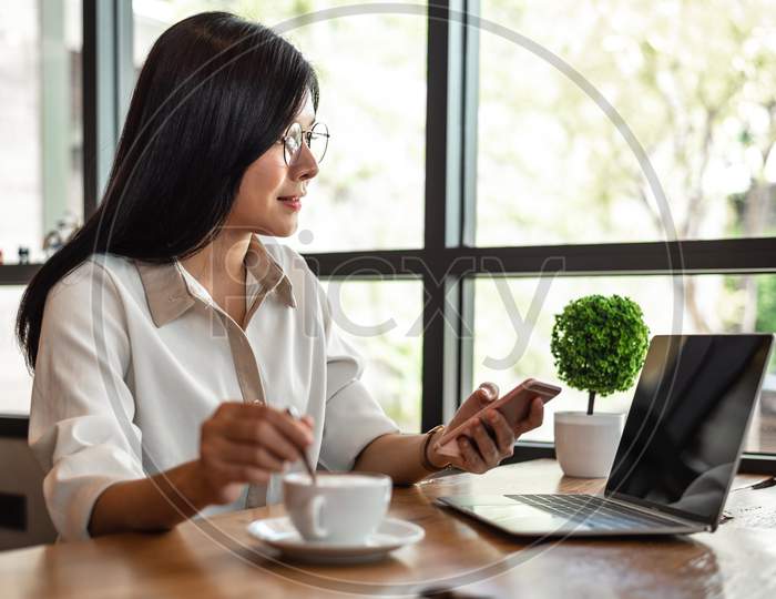 Business Woman Working With Laptop And Smartphone And Drinking Coffee In Office. Business And Lifestyles Concept. Entrepreneur And Freelance Theme. Selective Focus On Coffee Cup