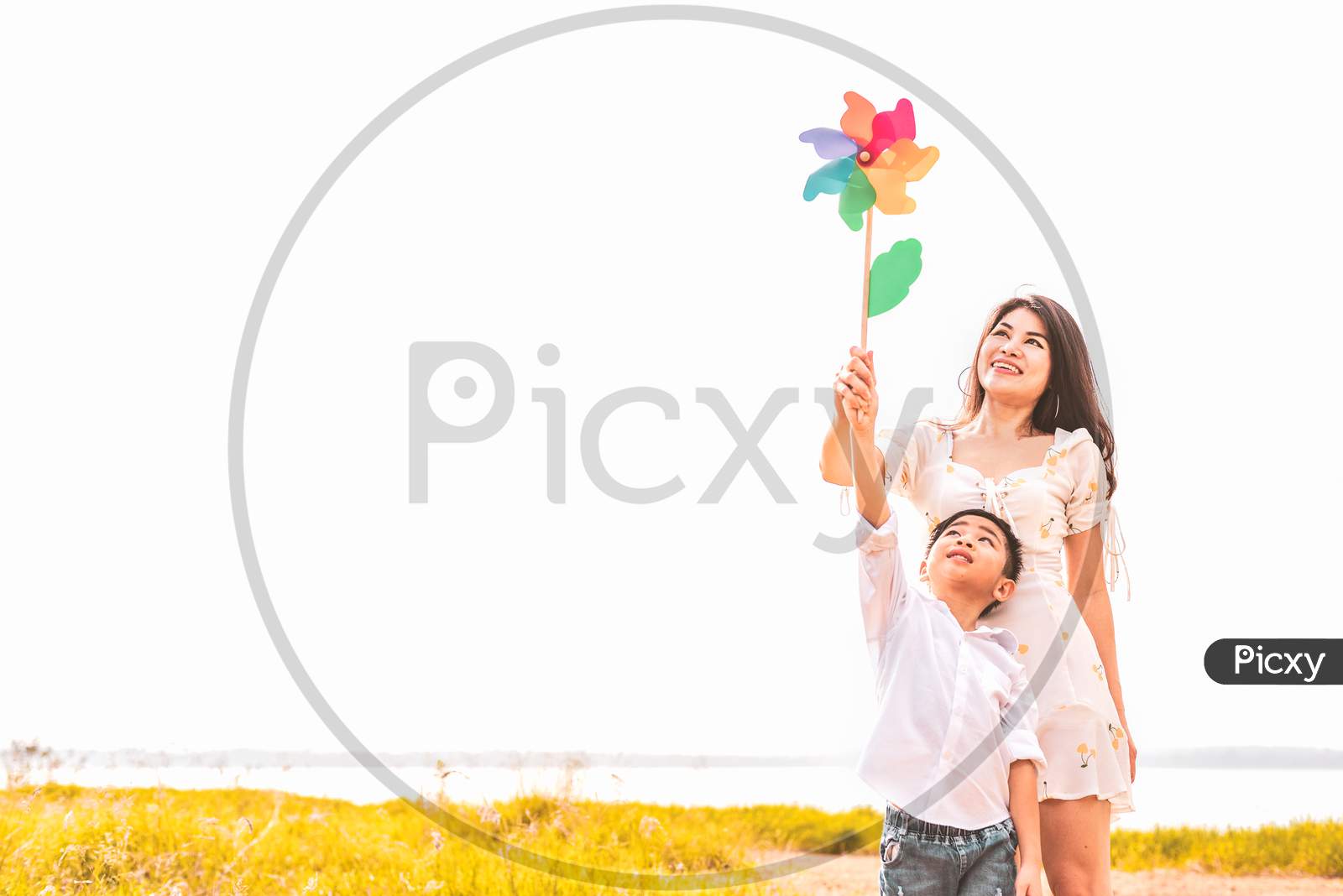 Little Asian Boy And His Mom Playing Colorful Rainbow Turbine In Meadow. Mother And Son Playing Together. Celebrating In Mother Day And Appreciating Concept. Summer People And Lifestyle Theme.